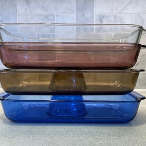 May include: Three glass baking dishes stacked on top of each other. The top dish is clear glass, the middle dish is brown glass, and the bottom dish is blue glass.