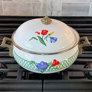 May include: White enamel pot with a floral design in red, blue, and yellow. The pot has a brass knob on the lid and brass handles. The pot is sitting on a black stovetop.