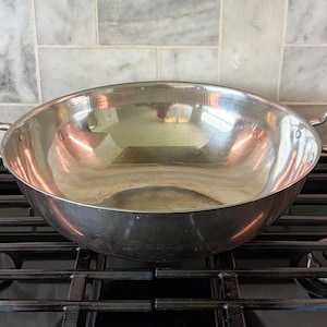 May include: A shiny, stainless steel wok-style pan with two curved handles. The pan sits on a black gas stovetop, reflecting the surrounding environment. The pan's interior is smooth and reflective.