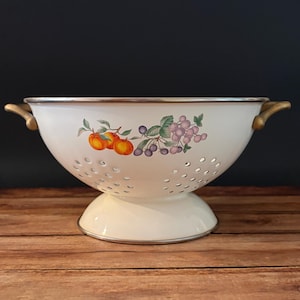 May include: A white enameled metal colander featuring a pedestal base, two gold-toned side handles, and a decorative fruit illustration of orange peaches and purple grapes with green leaves centered on the exterior bowl surface.