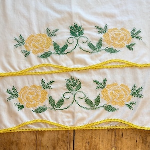 May include: Two white linen towels with yellow rose embroidery and green leaves. The towels have a yellow crocheted trim along the edges. The embroidery is centered on the towels.