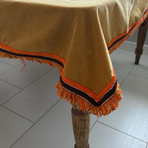 May include: A mustard-colored tablecloth with orange and brown trim and fringe. The tablecloth is draped over a wooden table, showcasing its textured fabric and decorative edges. The fringe adds a vintage touch.