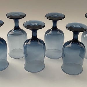 May include: Six blue-tinted wine glasses with a distinctive design. Each glass features a rounded bowl and a dark blue stem and base. The glasses are arranged in a row, showcasing their elegant shape and color.