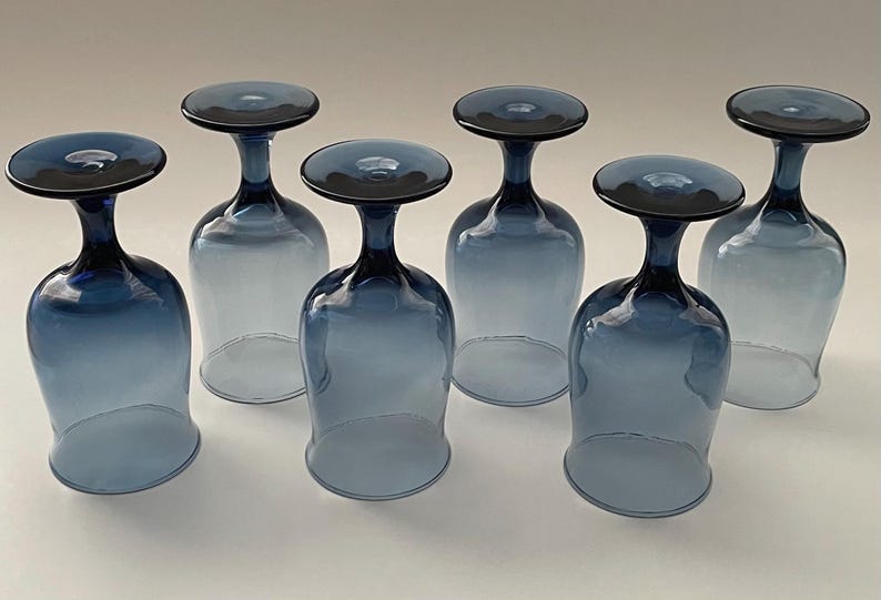 May include: Six blue-tinted wine glasses with a distinctive design. Each glass features a rounded bowl and a dark blue stem and base. The glasses are arranged in a row, showcasing their elegant shape and color.
