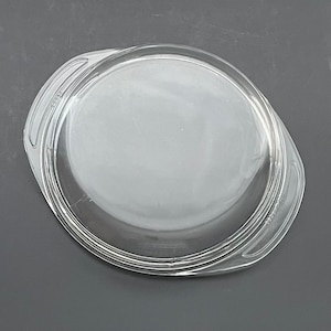 May include: A clear, round glass lid with two curved handles. The lid has a slightly raised rim and is designed to fit a round dish. The glass is transparent, allowing visibility of the contents. The lid is likely for a casserole dish or similar cookware.