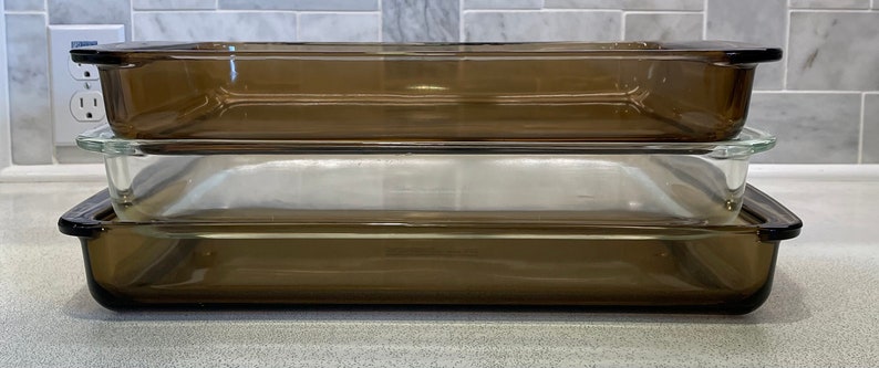 May include: Three rectangular glass baking dishes stacked on top of each other. Two are a transparent brown color, and one is clear. The dishes have a simple, functional design with slightly raised edges and handles.