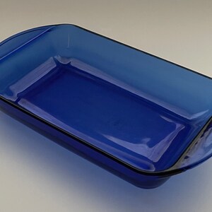 May include: A rectangular, cobalt blue glass baking dish with rounded corners and handles. The dish has a glossy finish and a black trim around the edges. This oven-safe dish is ideal for baking casseroles and other dishes.