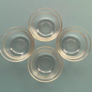 May include: Four clear glass bowls with a circular design. Each bowl has a slightly flared rim and concentric rings around the outside. The bowls are arranged in a square formation against a light blue background.