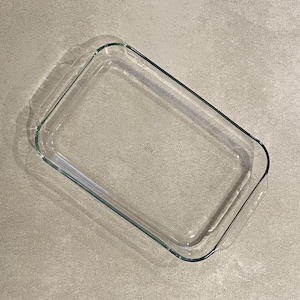 May include: A clear, rectangular glass baking dish with rounded corners. The dish is empty and sits on a light gray surface. The glass is transparent, allowing a view through the dish.