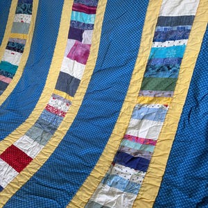 May include: A handmade quilt with a blue background and yellow borders. Vertical strips of various fabric patterns and colors, including red, white, and blue, are sewn together. The quilt has a soft, textured appearance, perfect for a bed or sofa.