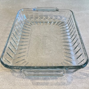 May include: A clear, rectangular glass baking dish with a ridged design on the sides and a smooth bottom. The dish has handles on each end and is designed for oven use. The Anchor Hocking brand name is visible.