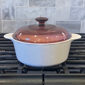 May include: A white ceramic casserole dish with a glass lid. The lid is a deep purple color and has a knob on top. The dish is sitting on a black stovetop.