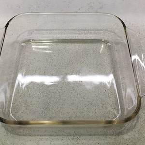 May include: A clear, square glass baking dish with rounded corners and two side handles. The dish is empty and sits on a speckled gray surface. Ideal for baking casseroles, desserts, or other dishes.