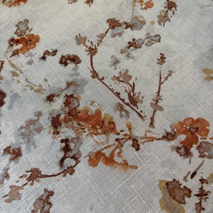 May include: Close-up of a light-colored fabric with a floral pattern. The design features branches and flowers in shades of brown, orange, and gray. The fabric has a textured appearance, with a subtle grid-like pattern visible throughout.