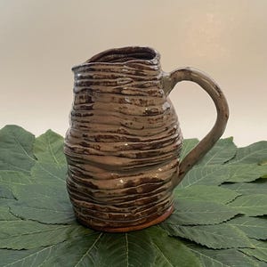 May include: A handmade brown ceramic pitcher with a textured, ridged surface. The pitcher has a curved handle and a spout for pouring. The pitcher is displayed on a bed of green leaves, creating a rustic and natural aesthetic.