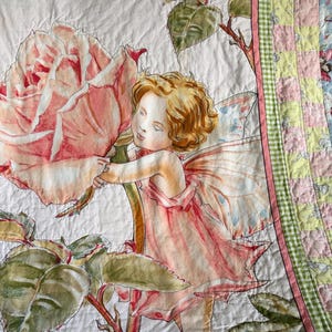 May include: A detailed illustration on a quilt features a fairy with curly blonde hair and pink wings, dressed in a pink dress, gently touching a large pink rose. The quilt has a patchwork border with green and pink accents.
