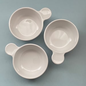 May include: Three white ceramic bowls with rounded handles. The bowls are empty and have a smooth, glossy finish.