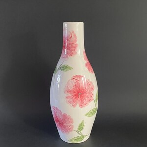 May include: A white ceramic vase with a slender neck and rounded body. The vase is decorated with pink floral designs and green leaves. The vase is approximately 10 inches tall.