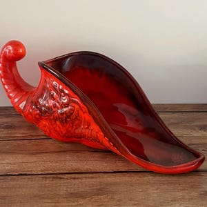 May include: A vibrant red ceramic decorative bowl with a unique, elongated shape. The bowl features a glossy finish and a textured, ornate design on the side. The bowl has a curved handle and is placed on a wooden surface.