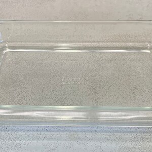 May include: A rectangular, clear glass baking dish with rounded corners. The dish is empty and sits on a speckled gray surface. The glass is transparent, allowing a view through the dish.