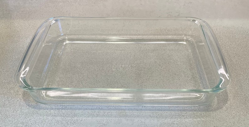 May include: A rectangular, clear glass baking dish with rounded corners. The dish is empty and sits on a speckled gray surface. The glass is transparent, allowing a view through the dish.