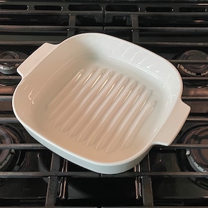 May include: A white ceramic grill pan with a rectangular shape and ridged interior. The pan has two handles and is set on a black stovetop. The pan is designed for grilling food.
