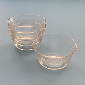 May include: A set of four clear glass bowls, stacked and arranged on a light blue surface. The bowls have a simple, rounded design with a slightly flared rim and horizontal ridges near the base. One bowl is separate, next to the stack.
