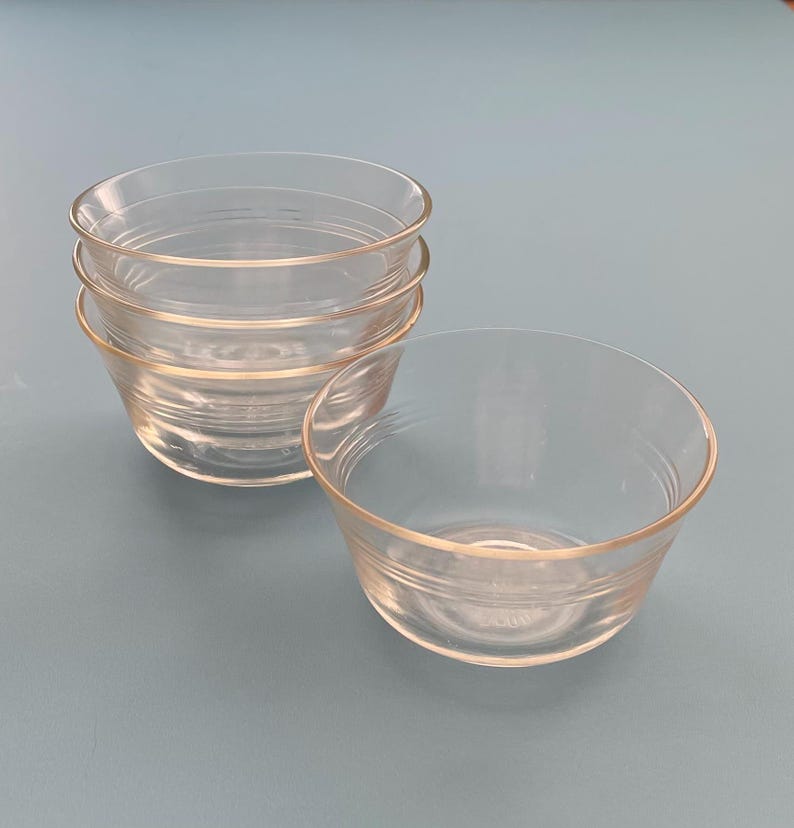 May include: A set of four clear glass bowls, stacked and arranged on a light blue surface. The bowls have a simple, rounded design with a slightly flared rim and horizontal ridges near the base. One bowl is separate, next to the stack.
