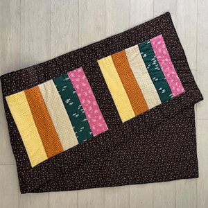 May include: A handmade quilt featuring two rectangular panels with vertical stripes in yellow, orange, cream, green, and pink. The quilt has a dark brown background with small white dots and a matching border. The quilt is folded.