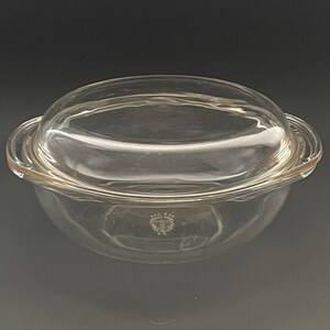 May include: A clear glass casserole dish with a matching lid. The dish has a rounded shape with a slight lip and two handles. The lid is domed. A decorative emblem is visible on the dish.