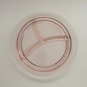 May include: A round, divided plate in a soft pink hue. The plate features three compartments, separated by curved dividers. The rim of the plate has a slightly raised edge. The plate is made of glass and is suitable for serving food.