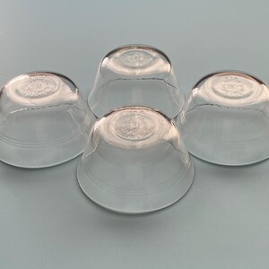 May include: Four clear glass bowls with a rounded shape and a slightly flared rim. Each bowl has a circular design on the bottom and two horizontal lines near the base. The bowls are arranged on a light blue surface.