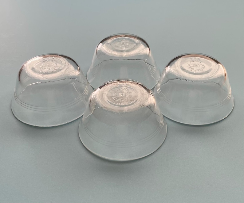 May include: Four clear glass bowls with a rounded shape and a slightly flared rim. Each bowl has a circular design on the bottom and two horizontal lines near the base. The bowls are arranged on a light blue surface.