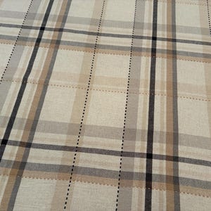 May include: A woven fabric featuring a plaid pattern in shades of beige, tan, gray, and black. The design consists of intersecting horizontal and vertical lines of varying widths, including thin dashed black lines that create a grid effect.