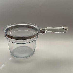 May include: A clear glass saucepan with a transparent handle and a silver-colored metal band around the top. The handle is attached to the pan with metal hardware. The pan is designed for cooking and has a simple, functional design.