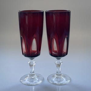 May include: Two ruby red champagne flutes. The tall, elegant glasses have a clear, ornate stem and base. The flutes have a vertical, cut design on the sides. The glasses are set against a neutral background.
