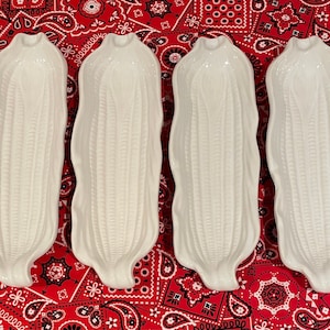 May include: Four white ceramic corn-shaped serving dishes. Each dish has a detailed, textured surface mimicking corn kernels. The dishes are arranged on a red paisley patterned fabric.