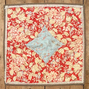 May include: A square, handmade quilt with a vibrant floral pattern in red, cream, and light blue. The quilt features a central diamond-shaped patch in light blue with floral details. The edges are finished with a lace trim and a brown border.