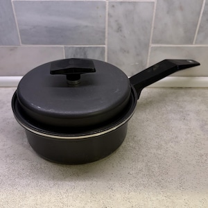 May include: A black saucepan with a lid and a long handle. The pan is sitting on a speckled countertop. The lid has a black handle. The pan is made of metal and has a silver rim.