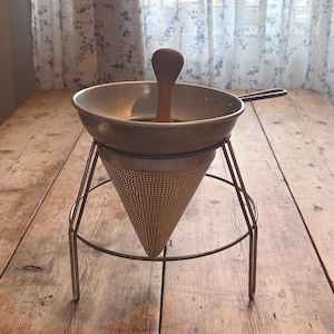 May include: A metal food mill with a conical perforated strainer, a wide circular rim, and a wooden pestle. The device is supported by a three-legged wire stand, featuring a side handle and a metallic finish with a natural wood handle on the pestle.