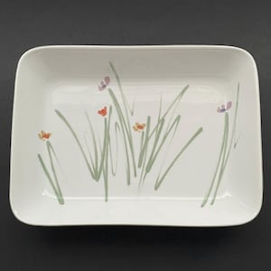 May include: A rectangular white ceramic serving dish with a floral design. The dish features hand-painted green grass and small orange and purple flowers. The dish has rounded corners and a smooth surface.