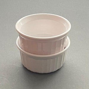 May include: Two white ceramic ramekins stacked on top of each other. The ramekins have a round shape with a slightly flared rim and vertical ridged detailing on the exterior. They are set against a gray background.