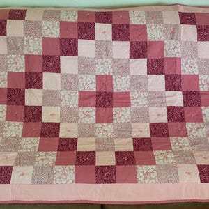 May include: A handmade quilt in shades of pink and white, featuring a patchwork design of squares. The quilt has a light pink border and is draped over a brown surface. The quilt's dimensions are approximately 70 inches by 80 inches.
