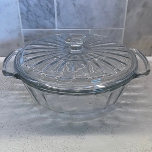 May include: A clear glass casserole dish with a matching lid. The lid has a ridged design and a small square handle. The dish has two handles and a geometric pattern on the sides. The dish is sitting on a white surface.
