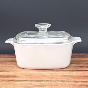 May include: A rectangular white casserole dish with integrated side handles and a clear glass lid featuring a central knob handle. The dish has smooth, rounded corners and a solid white finish, providing a clean and functional kitchen vessel.