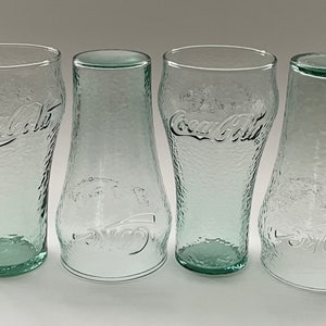 May include: Four clear Coca-Cola glasses with a textured surface. Two are the classic shape, and two are a unique bottle shape. The Coca-Cola logo is embossed on each glass. The glasses are made of clear glass with a slight green tint.