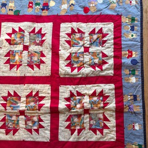 May include: A handmade quilt with a light blue border featuring teddy bears holding balloons. The quilt has red starburst patterns and squares with teddy bear illustrations in various outfits. The quilt is a patchwork design.