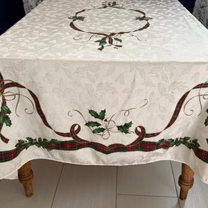 May include: Cream-colored tablecloth with a repeating leaf pattern. The edges are decorated with red and green plaid ribbons, holly, and bows. A matching design is centered on the tablecloth. The tablecloth is draped over a wooden table.