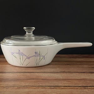 May include: White casserole dish with a clear glass lid and a long handle. The dish is decorated with a floral iris design in purple and green. The dish is on a wooden surface.