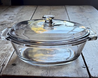 May include: A clear glass casserole dish with a matching lid. The round vessel features two integrated side handles and a central knob on the lid. The glass is transparent, allowing a view through the bowl and the lid, which sits securely on top.